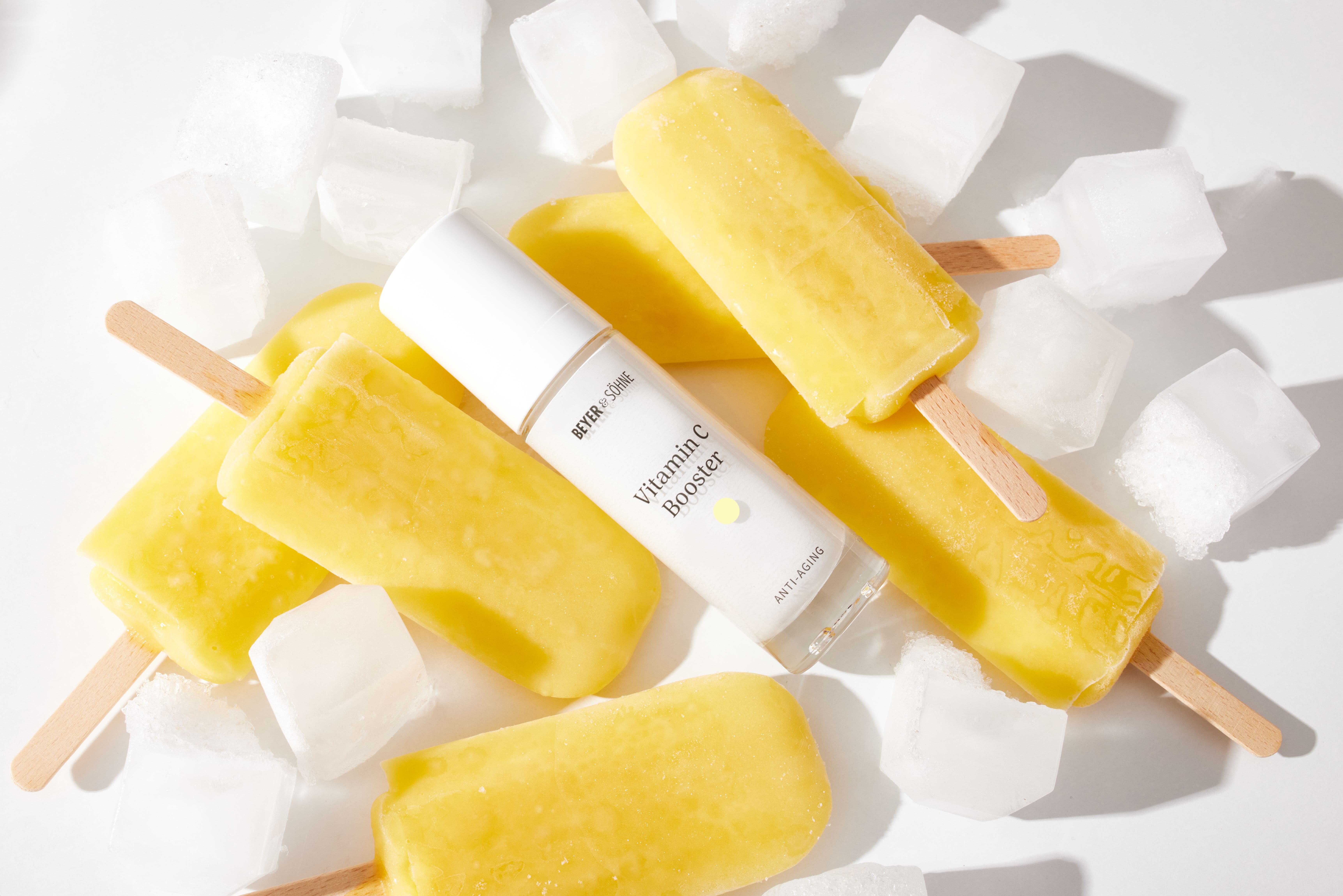 Vitamin C Booster von Beyer & Söhne in länglicher, weißer Glasflasche liegt zwischen gelbem Wassereis und Eiswürfeln und vermittelt Frische